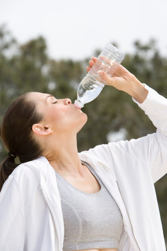 Woman in athletic wear drinking hydrogen water from a plastic bottle outdoors, with trees blurred in the background.