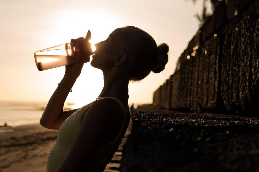 Silhouetted person drinking from a bottle at sunset on a beach, leaning against a textured wall.