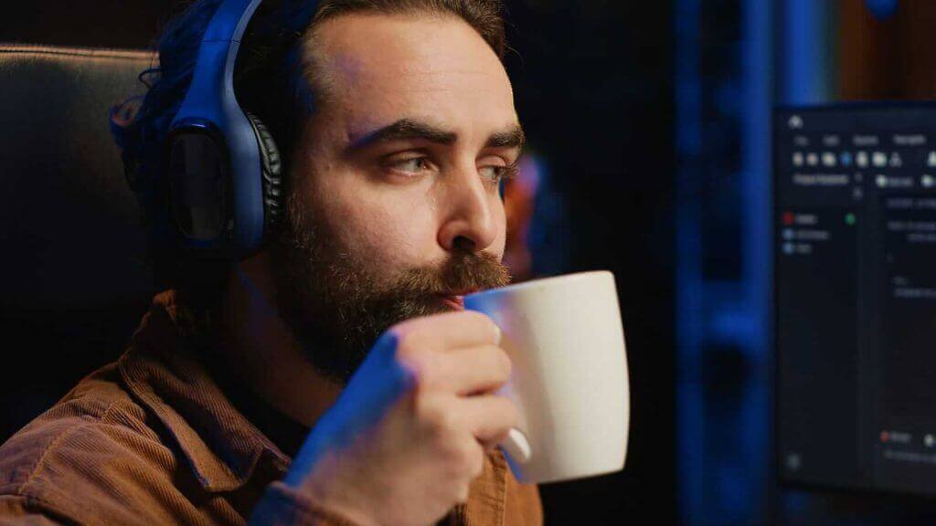 A person wearing headphones sits at a desk, sipping from a white mug while looking at a computer screen.