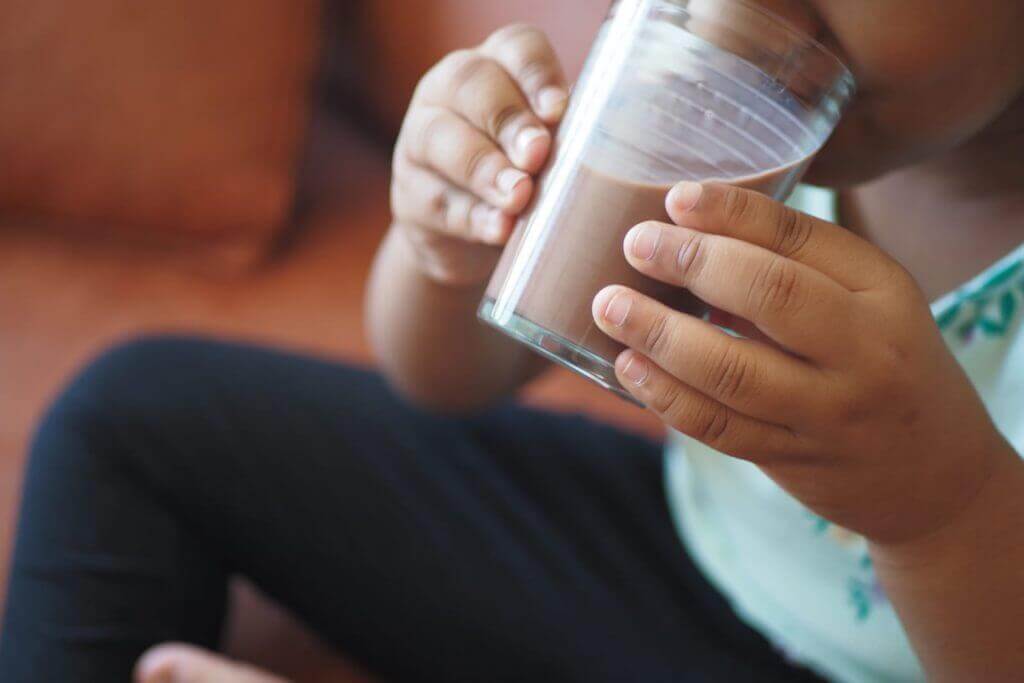 Child holding a clear glass of chocolate milk while sitting on a brown couch, wearing a light-colored shirt and dark pants.