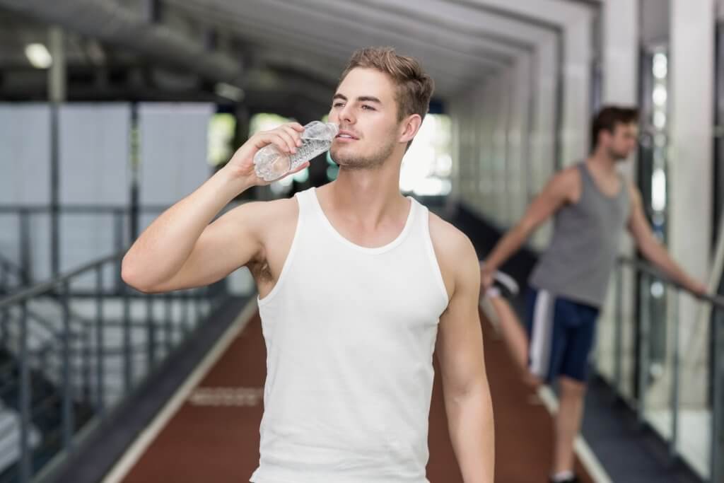 Man in a white tank top drinking water from a bottle in an indoor athletic facility. Another person is stretching in the background.