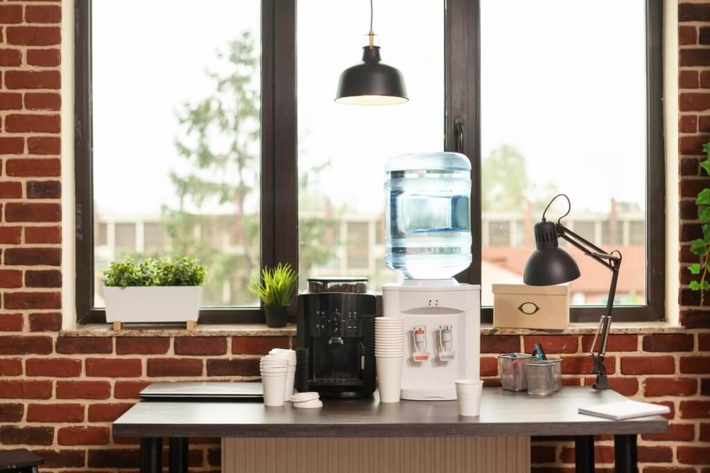 Office break area with a water cooler, coffee machine, and desk lamp. A row of disposable cups is on the table, which is set against a brick wall with two large windows.