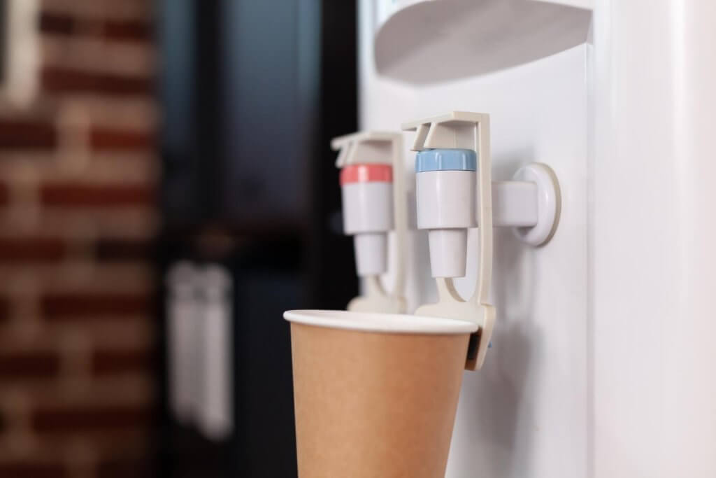 A paper cup positioned under a water dispenser with two taps, one red for hot water and one blue for cold water.