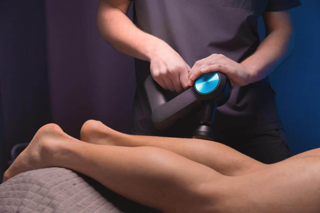 Person using a massage gun on someone's legs in a dimly lit room.