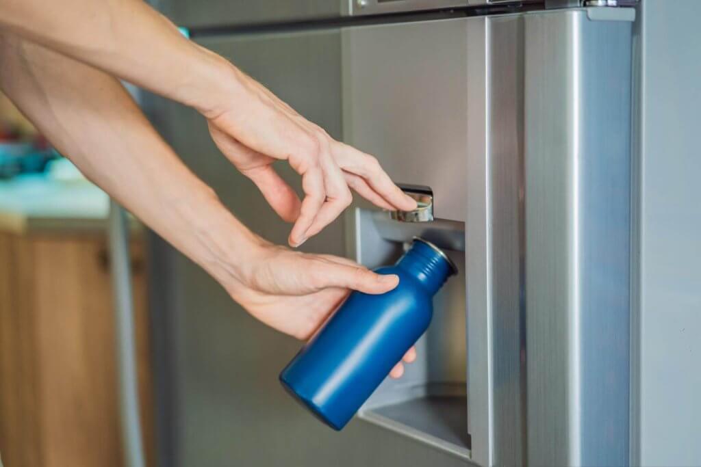 A person fills a blue water bottle at a refrigerator water dispenser.