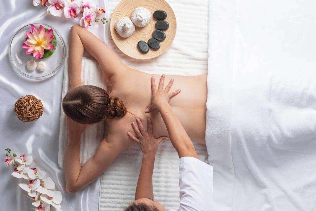 Person receiving a back massage on a white massage table, surrounded by orchids, candles, and stones.