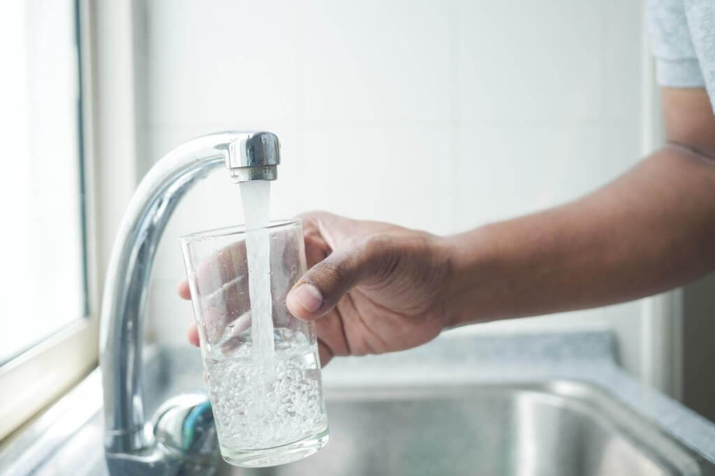 A person filling a glass with water from a kitchen faucet over a sink.