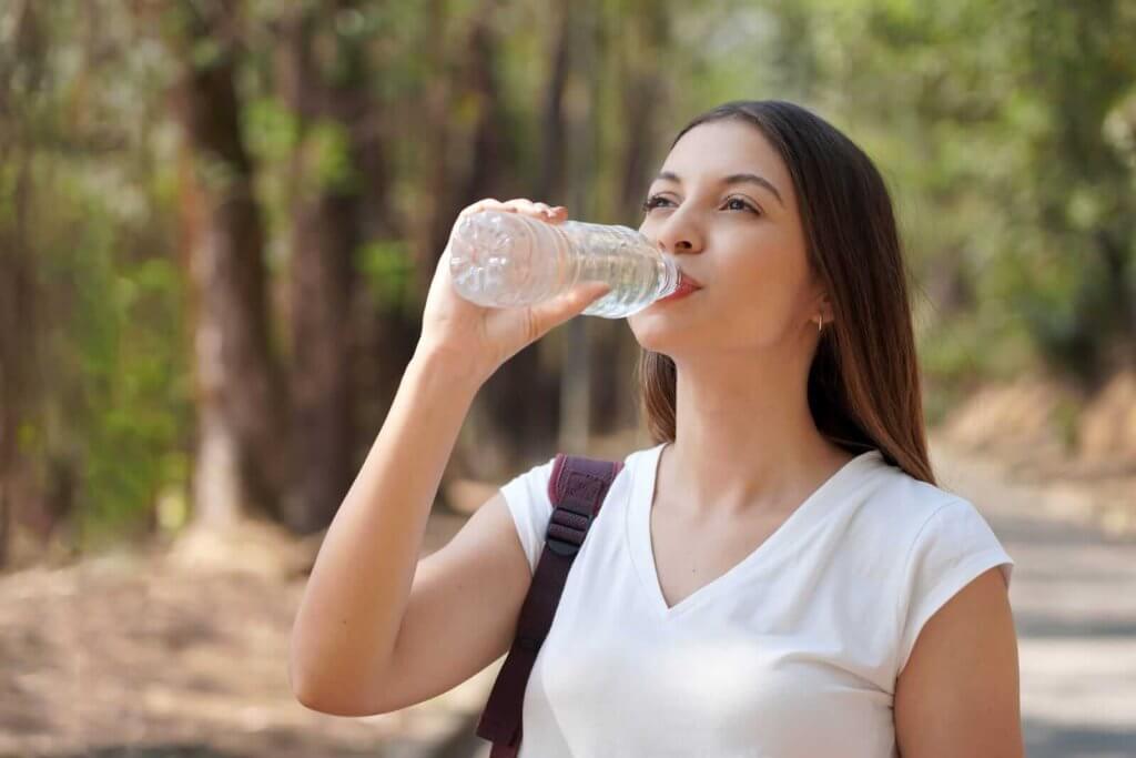 A woman in a white shirt drinks from a plastic water bottle while standing on a path surrounded by trees.