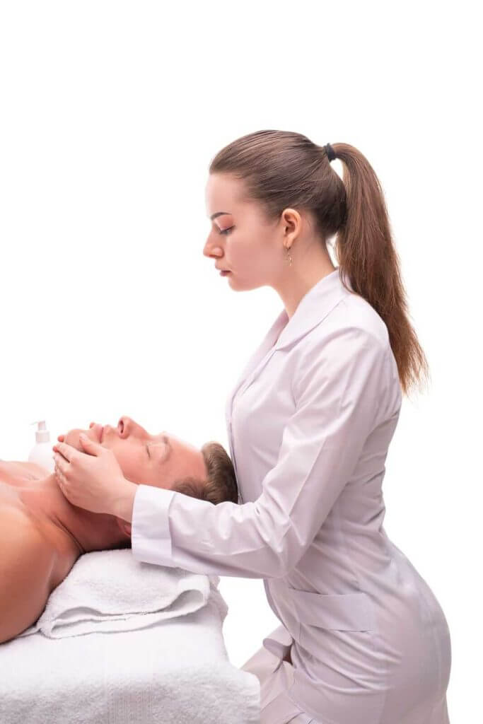 A woman in a white coat performs a facial massage on a man lying on a treatment bed.