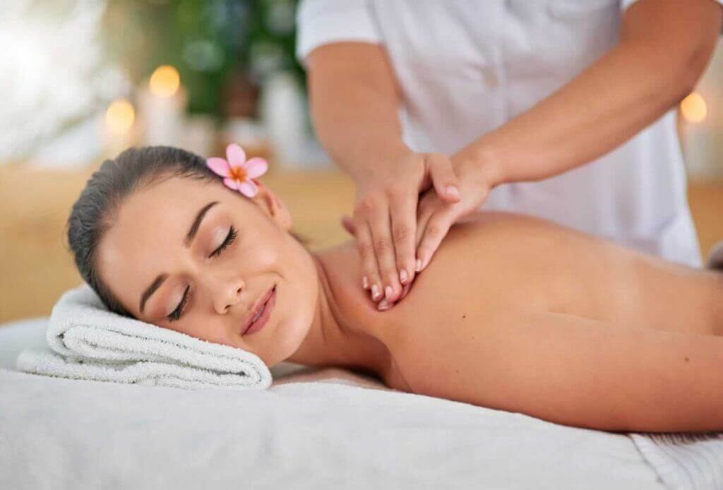 A woman receiving a back massage while lying on a white towel, with a flower in her hair.