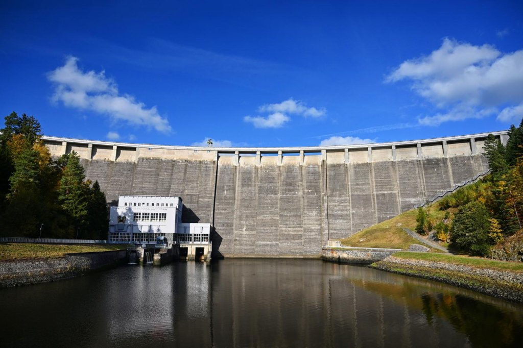A large concrete dam with a hydroelectric power station at its base, surrounded by trees and reflecting in a calm reservoir under a blue sky.