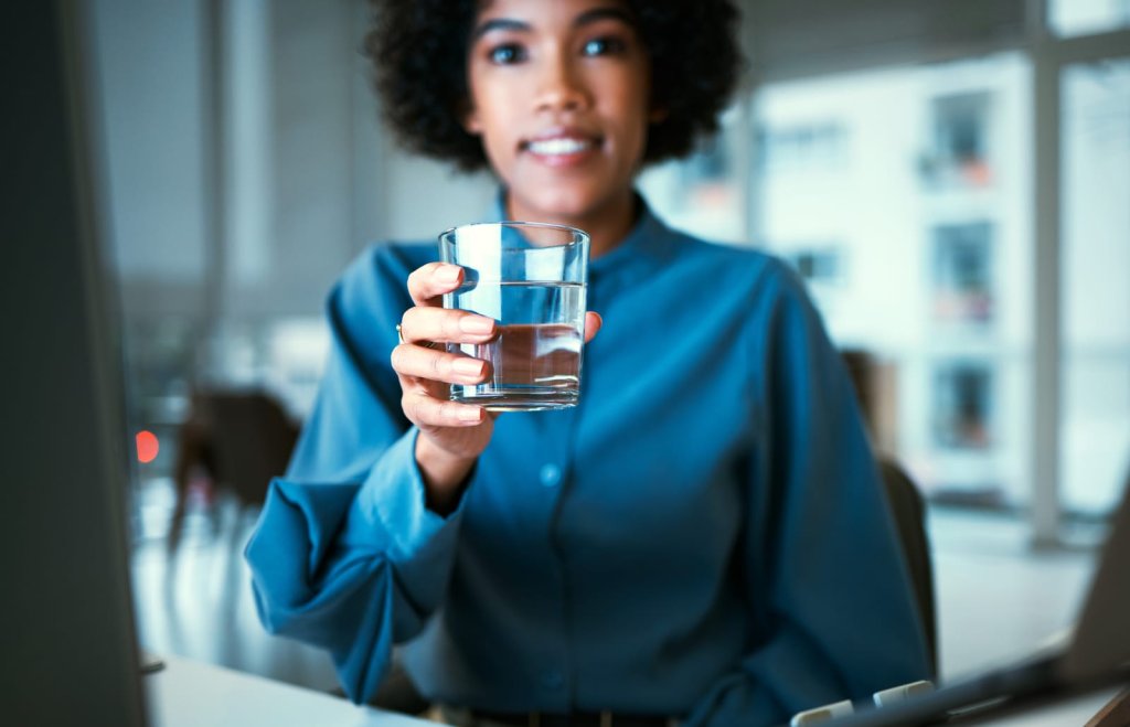 Person wearing a blue shirt holding a glass of water while sitting at a desk in a bright office environment.