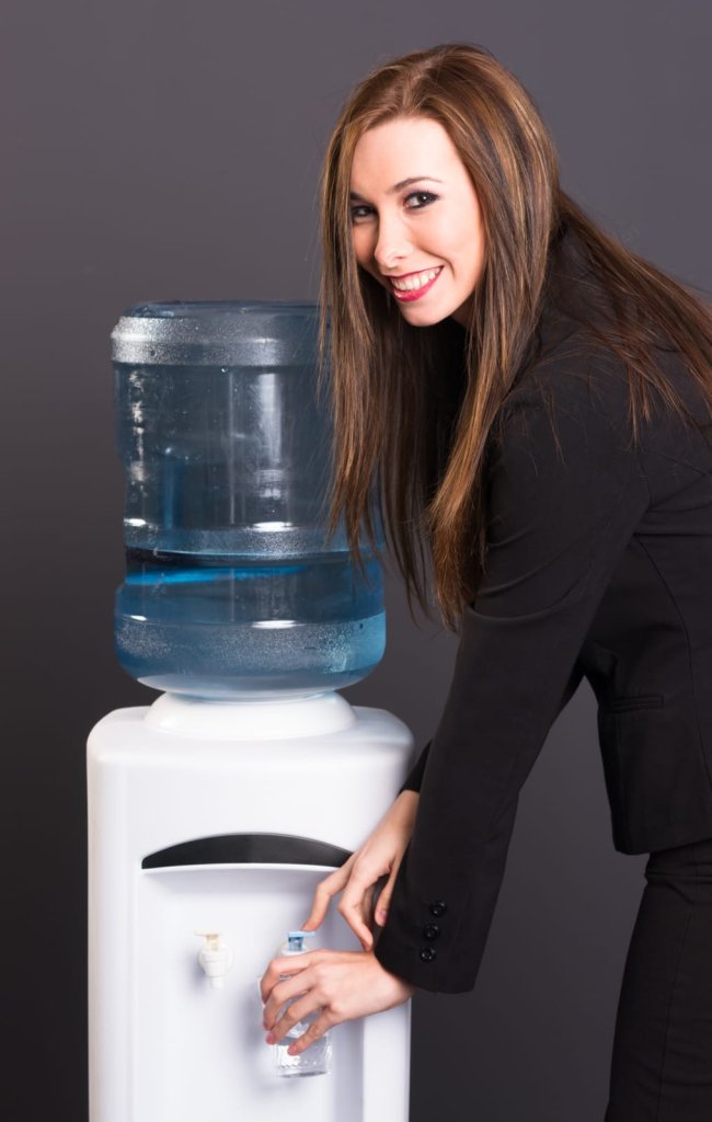 A woman in a black suit fills a plastic cup with water from a water cooler against a plain dark background.