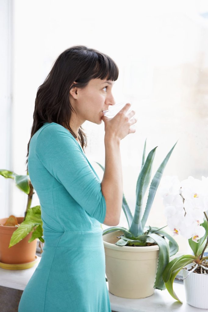 A woman in a teal dress drinks water while standing by a window with potted plants.
