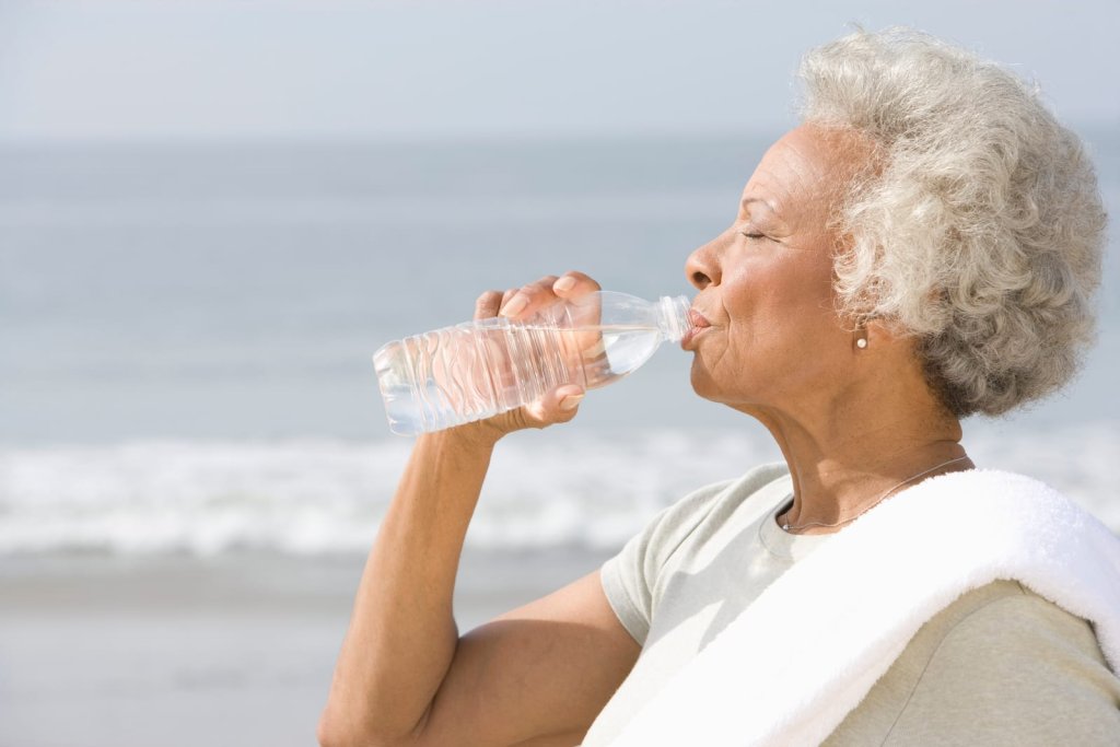 Older woman drinking water from a plastic bottle at the beach, with a towel over her shoulder and the ocean in the background.