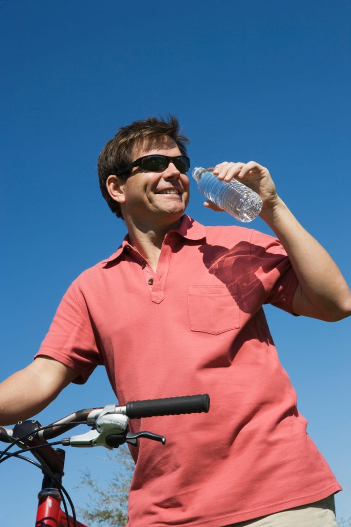 Man wearing sunglasses and a pink polo shirt stands outdoors with a bicycle, holding a water bottle and smiling under a clear blue sky.