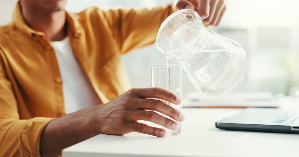 A person pours water from a glass pitcher into a glass while sitting at a table with a laptop.