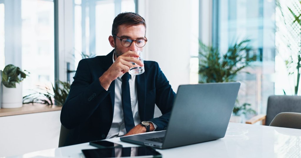 A man in a suit sits at a desk, drinking water while working on a laptop in a modern office setting.