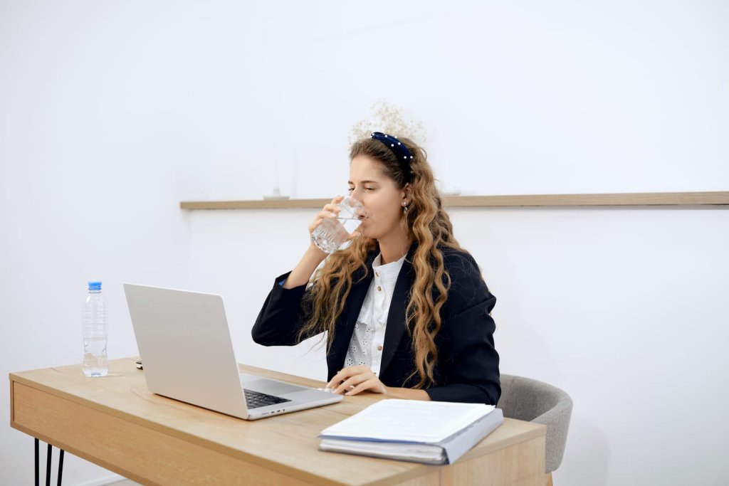 A woman sits at a desk with a laptop and a notebook, drinking water from a glass.