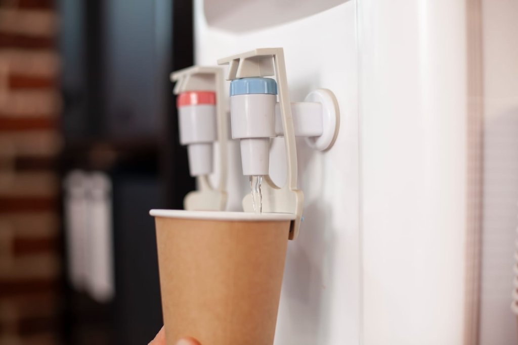 A paper cup is held under the cold water tap of a white water dispenser as water pours into the cup, highlighting convenience and affordability similar to top water dispenser Singapore price options.