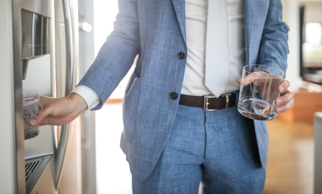 A person in a blue suit fills a glass with water from a refrigerator dispenser while holding another empty glass, possibly considering the water dispenser Singapore price for their next purchase.