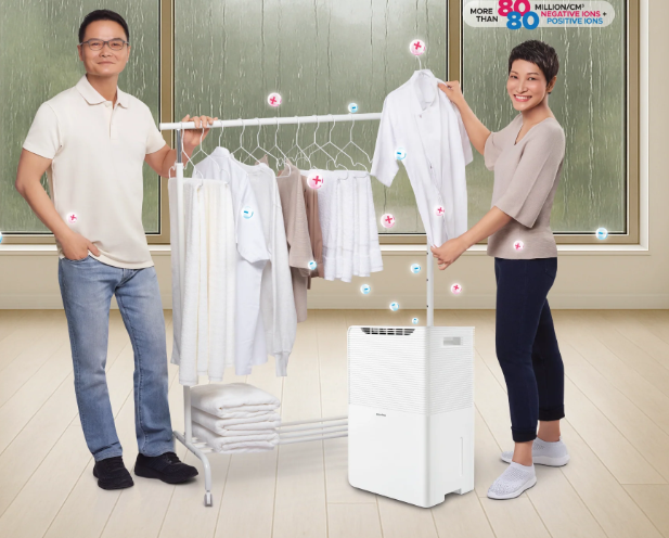Two people stand beside a drying rack with clothes and a white dehumidifier indoors, with rain visible through a window in the background. Blue and red icons appear over the clothes, highlighting how dehumidifiers can help keep laundry dry even on rainy days.
